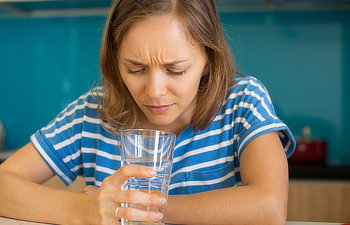 dissatisfied young beautiful woman looking into glass of water and sitting at table in kitchen