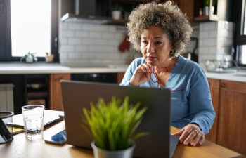 A concerned mature woman with curly gray hair reviews paperwork at her kitchen table. She holds a document while looking at it intently, surrounded by a laptop, calculator, and papers