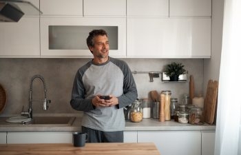A casually dressed man stands in a bright, contemporary kitchen, looking at his phone with a smile. The counter features jars, cutting boards, and kitchen tools, suggesting everyday home life.