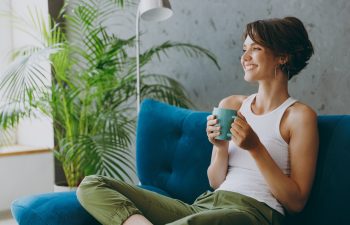 Side view young woman wears white tank shirt drink tea coffee sit on blue sofa couch stay at home hotel flat rest relax spend free spare time in living room indoors grey wall. People lounge concept